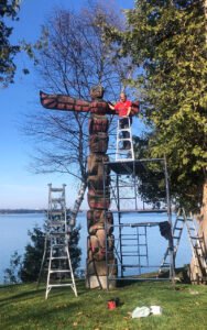 Artist works on a totem pole by a lakeside in a recent project
