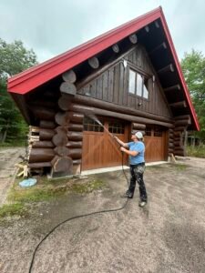 Person power washing log cabin with red roof in forest